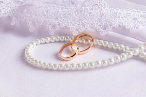 Two gold wedding rings lie on a string of pearl beads. on a white satin background with an embroidered veil. The concept of the wedding. Front view. photo