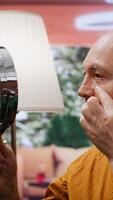 Vertical Senior man looking at his aging face in the mirror and trying to preserve his skin, using hydration and UV protection serum. Elderly person applying grooming products on aging skin. Camera B. video