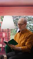 Vertical Elderly man reading a book sitting alone in his lounge area at home, relaxing and feeling good with free time. Person with an intellectual hobby, passionate about literature and lecture. Camera B. video