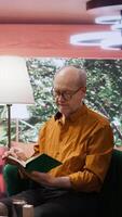 Vertical Senior person enjoying lecture with a book and relaxing on couch at home, leisure activity to train his imagination during retirement. Old man reading literature as a pastime hobby. Camera B. video