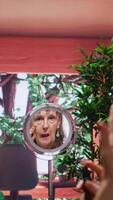 Vertical Senior woman looking in the mirror and applying anti aging cream on her face, using rejuvenating products to preserve her skin. Pensioner doing pampering routine with serum and lotion. Camera B. video