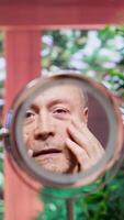 Vertical Pensioner using serum and cream to preserve his mature skin with wrinkles, looking in the mirror at his aging face. Doing a pampering daily routine for personal care, grooming products. Camera A. video