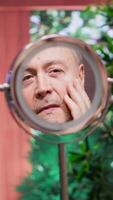 Vertical Elderly man looking in the mirror at his wrinkles and aging skin while he does his daily routine for grooming and skincare. Applying rejuvenating cream and lotion for hydration. Camera A. video