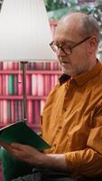 Vertical Elderly man reading a book sitting alone in his lounge area at home, relaxing and feeling good with free time. Person with an intellectual hobby, passionate about literature and lecture. Camera A. video