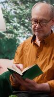 Vertical Senior person enjoying lecture with a book and relaxing on couch at home, leisure activity to train his imagination during retirement. Old man reading literature as a pastime hobby. Camera A. video