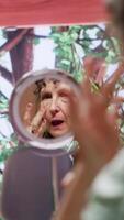 Vertical Senior woman looking in the mirror and applying anti aging cream on her face, using rejuvenating products to preserve her skin. Pensioner doing pampering routine with serum and lotion. Camera A. video