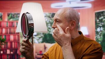 Senior man looking at his aging face in the mirror and trying to preserve his skin, using hydration and UV protection serum. Elderly person applying grooming products on aging skin. Camera B. video