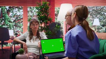 Medical assistant holding a laptop with isolated copy space display, consulting elderly woman in wheelchair during a social home visit. Nurse offering health aid support and advice. Camera B. video