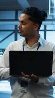 Vertical Technician using laptop in data center, managing servers, operating systems. Professional in server hub using notebook to ensure networks are secure and functioning properly, camera A video