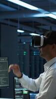 Vertical Admin in data center using virtual reality tech to ensure system can handle networking tasks. Server room worker wearing VR headset to track network traffic with PC, making improvements, camera A video