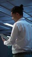 Vertical System administrator overseeing data center, making improvements and updates to ensure system can handle networking tasks. IT expert in server room tracking network traffic with tablet, camera B video