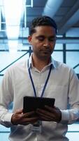 Vertical System administrator walks in data center used for managing gear energy consumption, working on tablet. Close up of IT specialist using device in workspace used for automation in server room, camera B video