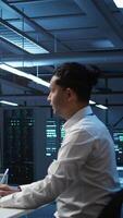 Vertical Team of programmers in high tech data center troubleshooting issues using tablet. Teamworking colleagues in server farm using device, establishing performance benchmarks, camera B video