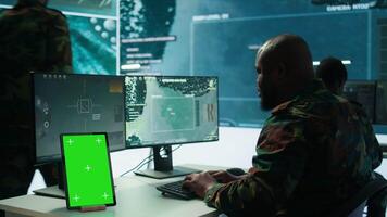 Soldier gathering real time surveillance data next to mockup display in high tech command center, processing intelligence for field operations. Working with modern gear and big screen. Camera B. video