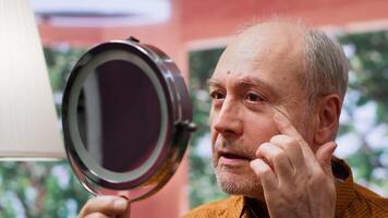 Senior man looking at his aging face in the mirror and trying to preserve his skin, using hydration and UV protection serum. Elderly person applying grooming products on aging skin. Camera A. video