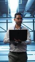Vertical Technician using laptop in data center, managing servers, operating systems. Professional in server hub using notebook to ensure networks are secure and functioning properly, camera B video