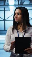Vertical Software developer walking in data center, using tablet to design, implement and manage gear, ensuring optimal performance and reliability. IT worker in server farm coding on device, camera B video