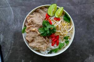 A Bowl Pho Bo traditional Soup with beef, rice noodles, ginger, lime, chili pepper in bowl. Close up. Vietnamese cuisine served on black background photo