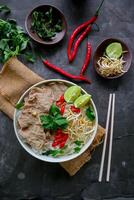 A Bowl Pho Bo traditional Soup with beef, rice noodles, ginger, lime, chili pepper in bowl. Close up. Vietnamese cuisine served on black background photo