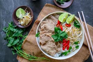 A Bowl Pho Bo traditional Soup with beef, rice noodles, ginger, lime, chili pepper in bowl. Close up. Vietnamese cuisine served on black background photo
