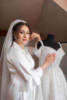 A woman is standing in front of a wedding dress, holding it up to her chest. The dress is white and has a lot of sparkles photo