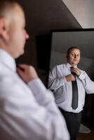 A man is getting ready for a formal event, adjusting his tie in front of a mirror. Concept of preparation and attention to detail, as the man carefully ties his tie to ensure it looks neat photo