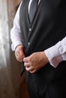 A man in a black vest and a white shirt is getting ready to dress up. He is adjusting his tie and putting on a ring. Concept of formality and preparation for a special occasion photo