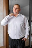 A man in a white shirt is standing in front of a mirror, adjusting his collar. Concept of formality and attention to detail, as the man takes care to ensure his appearance is neat and polished photo