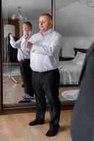 A man in a suit is standing in front of a mirror, adjusting his tie. Concept of formality and attention to detail, as the man takes the time to ensure his appearance is polished and professional photo
