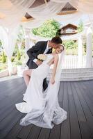 A bride and groom are posing for a picture in front of a pavilion. The bride is wearing a white dress and the groom is kissing her on the cheek. Scene is romantic and happy photo