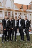 A group of five men in suits are posing for a photo in front of a building. Scene is formal and professional, as the men are dressed in suits and ties. The setting of the photo is a courtyard or park