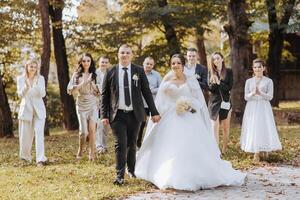 A bride and groom are walking down a path with a group of people behind them. The bride is wearing a white dress and the groom is wearing a suit. The group of people are all dressed up photo