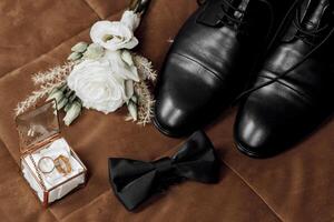 A man's wedding shoes and bow tie are displayed on a brown surface. The flowers are white and are placed in a small box photo