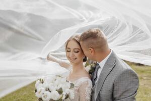 A bride and groom are standing in front of a white veil, with the bride holding a bouquet of flowers. Scene is romantic and happy, as the couple is embracing and enjoying their special day together photo