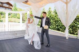 A bride and groom are dancing together in a garden. The bride is wearing a white dress and the groom is wearing a suit. They are both smiling and enjoying the moment photo