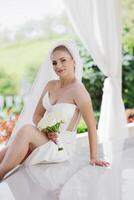 A bride is sitting on a table with a bouquet of flowers in her lap. She is wearing a white dress and a veil. The scene is set in a garden or a courtyard, with a white curtain in the background photo