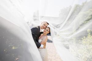 A bride and groom are posing for a picture under a white veil. The bride is wearing a white dress and the groom is wearing a gray suit. Scene is happy and romantic photo