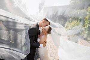 A bride and groom are embracing under a white veil. The bride is wearing a white dress and the groom is wearing a suit. The scene is intimate and romantic, capturing the love photo