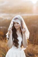 A woman in a white dress is standing in a field with a veil over her head. She is smiling and looking towards the camera. The scene has a peaceful and serene mood, with the woman's dress photo