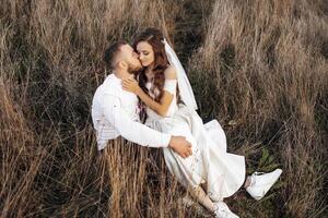 A bride and groom are sitting in a field, kissing each other. Scene is romantic and intimate photo