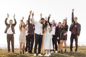 A group of people are posing for a picture, with a bride and groom in the center. The bride is wearing a white dress and the groom is wearing a suit. The group is standing on a grassy field photo