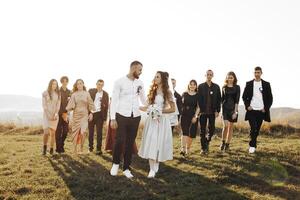 A bride and groom are walking down a grassy field with a group of people behind them. The bride is holding a bouquet and the groom is holding her hand. The atmosphere is joyful and celebratory photo