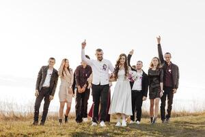 A group of people are standing in a field, with a bride and groom in the center. The bride is holding a bouquet and the groom is holding her hand. The group is all smiling and looking happy photo