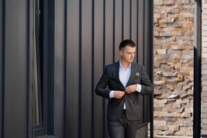 A man in a suit is standing in front of a brick wall. He is adjusting his jacket and looking at the camera photo