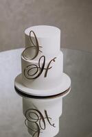 A white cake with the letters J and H on it. The cake is two layers and has a gold lettering photo