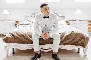 A man in a suit and bow tie sits on a bed. The bed is white and has a brown comforter. The man is looking at the camera photo