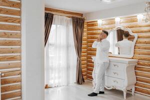 A man is getting ready in a room with a wooden floor and a wooden dresser. The room has a rustic feel to it, with wooden furniture and a window. The man is standing in front of a mirror photo