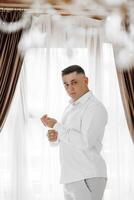 A man in a white shirt is standing in front of a window, adjusting his shirt. Concept of formality and attention to detail, as the man takes care to ensure his appearance is neat and tidy photo