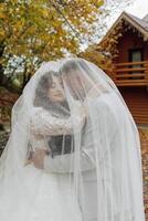 A bride and groom are hugging each other under a veil. The bride is wearing a white dress and the groom is wearing a suit. The scene is set in a forest, with trees in the background photo