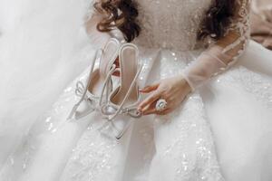 A woman is wearing a white dress and holding two pairs of shoes. The shoes are adorned with rhinestones and are tied with a bow. The woman is sitting on a bed photo